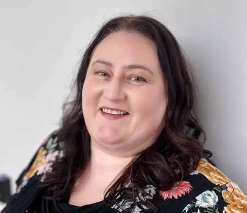 A smiling white woman with medium-length dark hair, wearing a dark floral shirt.