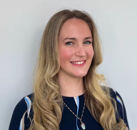 A smiling white woman with long blonde hair, wearing a dark blue patterned blouse.