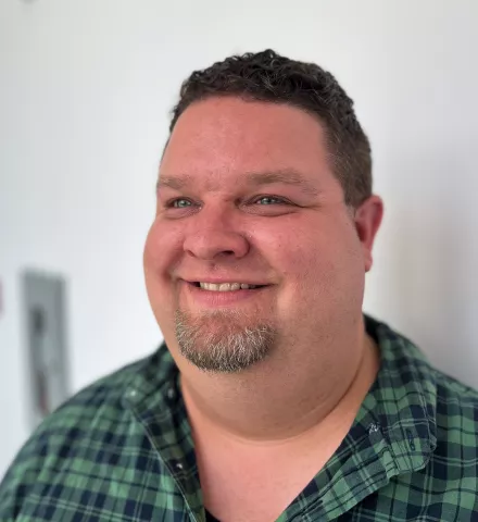 A photograph of a white man with short brown hair and a small beard, wearing a green and black plaid shirt.